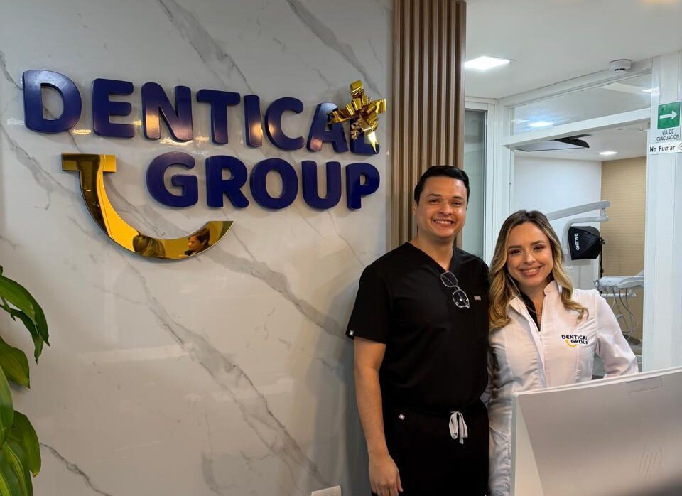 Dos dentistas sonríen en la recepción de una clínica dental con el logo "Dentica Group" en la pared.