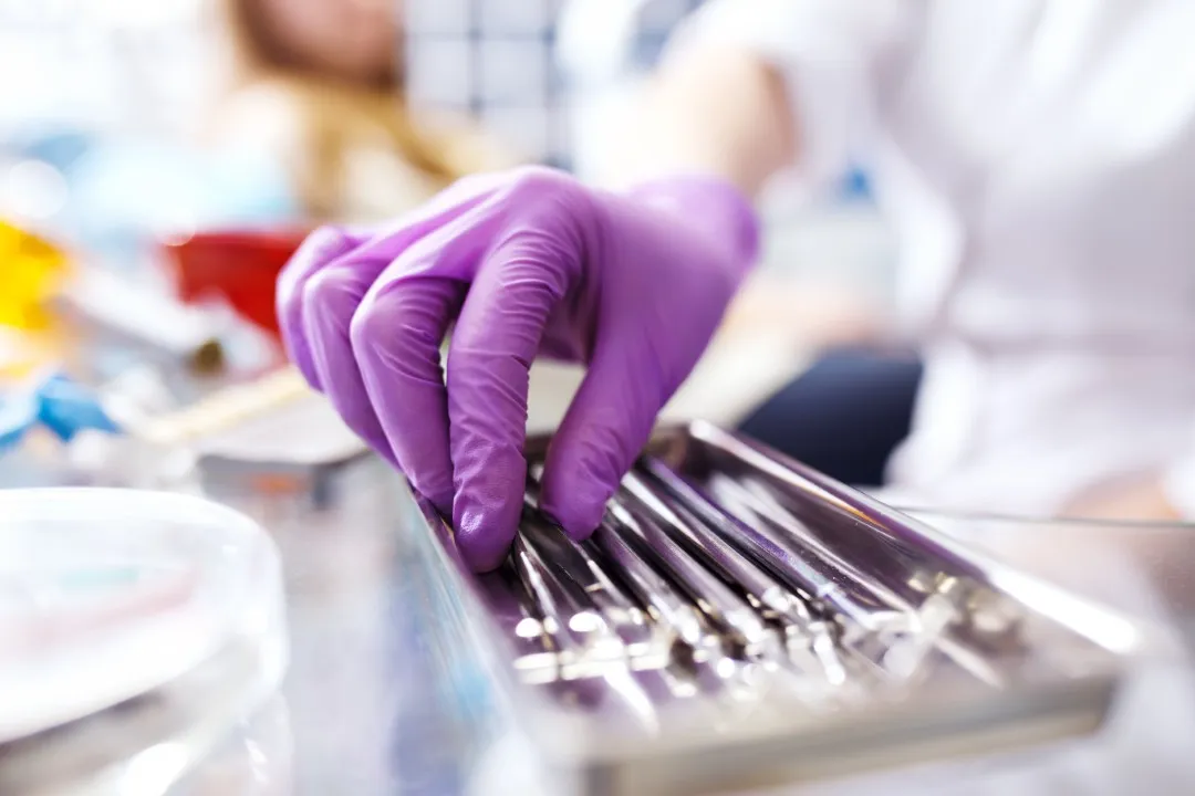 Una mano con guante morado toma instrumentos dentales de una bandeja metálica en un entorno de odontología general.